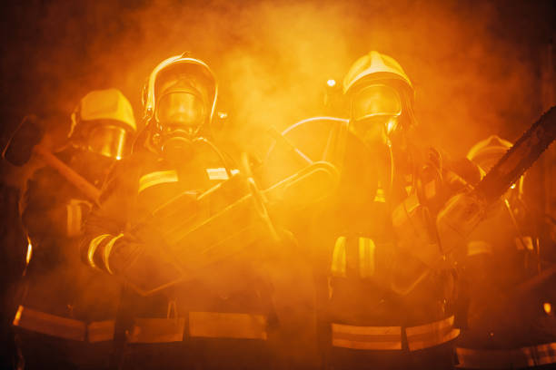Group of professional firefighters wearing full equipment, oxygen masks, and emergency rescue tools, circular hydraulic and gas saw, axe, and sledge hammer. smoke and fire trucks in the background. stock photo