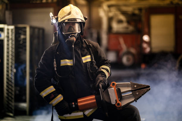 istock firefighter portrait wearing full equipment, oxygen mask, and power hydraulic cutting tool, smoke and fire trucks in the background. 1341135538