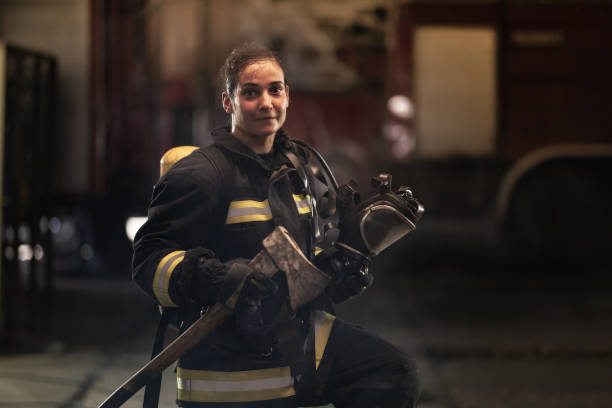 female firefighter portrait wearing full equipment, oxygen mask, and an axe. smoke and fire trucks in the background. stock photo