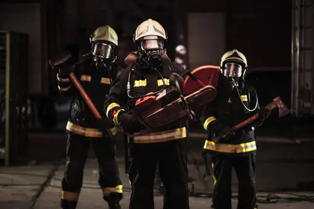 Group of professional firefighters wearing full equipment, oxygen masks, and emergency rescue tools, circular hydraulic and gas saw, axe, and sledge hammer. Firetrucks in the background.