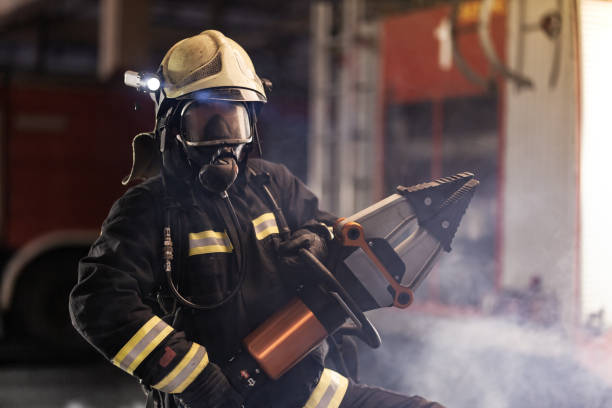 istock firefighter portrait wearing full equipment, oxygen mask, and power hydraulic cutting tool, smoke and fire trucks in the background. 1341136120