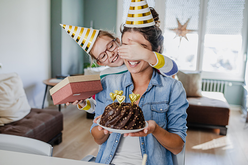 Young mother and daughter are at home, they are wearing party hats and holding a birthday cake