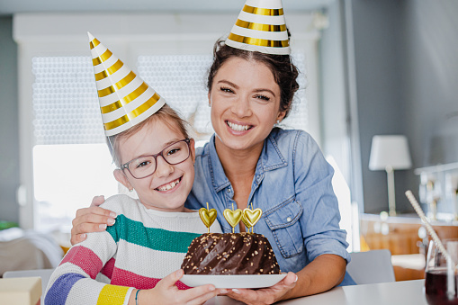 Young mother and daughter are at home, they are wearing party hats and holding a birthday cake