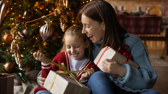Laughing sincere young woman cuddling curious happy kid daughter, unwrapping Christmas gifts together sitting near decorated festive tree in living room, magic winter holiday morning concept.