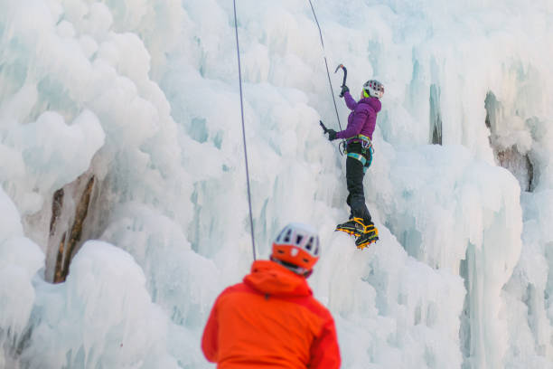 Safety measure during ice climbs Man standing and controlling a safety top rope while female with ice climbing equipment, climbing on a frozen waterfall Icefall stock pictures, royalty-free photos & images