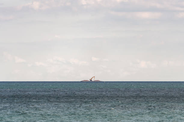 ein lastkahn mit baumaschinen und baggern mit steinhaufen auf beiden seiten schwimmt über das wasser des michigansees am horizont und wird von einem schlepper weit in der ferne gezogen. - industrial ship earth mover barge yellow stock-fotos und bilder