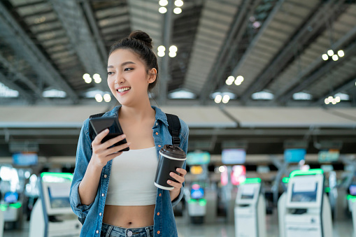 young attractive asian female traveller wearing face mask hand use smartphone checking map and flight schedule waking with luggage and handbag at airport terminal new normal travel lifestyle