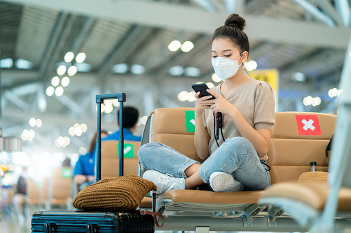 young attractive casual asian female adult woman wearing face mask sit relax hand use smartphone social media technology sit with social distancing at airport terminal waiting area