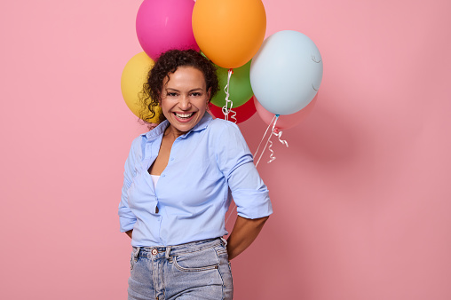 Charming joyful African woman in blue shirt laughing and smiling with toothy smile looking at the camera, holding multi-colored air balloons behind her back. Pink colored background, copy space