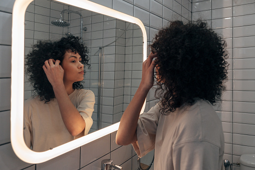 Joven y bonita revisándose en el espejo en el baño moderno. Poner el pelo rizado detrás de la oreja photo