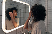 Young pretty woman checking herself in the mirror in modern bathroom. Putting curly hair behind ear