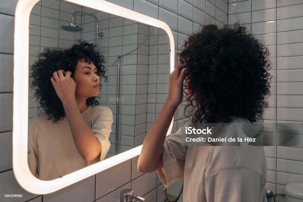 Young pretty woman checking herself in the mirror in modern bathroom. Putting curly hair behind ear Young pretty woman checking herself in the mirror in modern bathroom. Putting curly hair behind ear. Female selfcare concept. At home concept. Mirror - Object Stock Photo