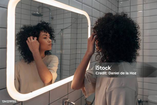 Young Pretty Woman Checking Herself In The Mirror In Modern Bathroom Putting Curly Hair Behind Ear Stock Photo - Download Image Now
