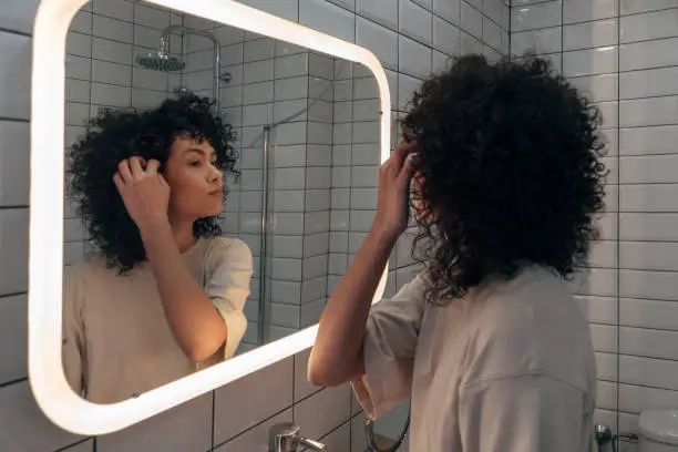 Young pretty woman checking herself in the mirror in modern bathroom. Putting curly hair behind ear. Female selfcare concept. At home concept.