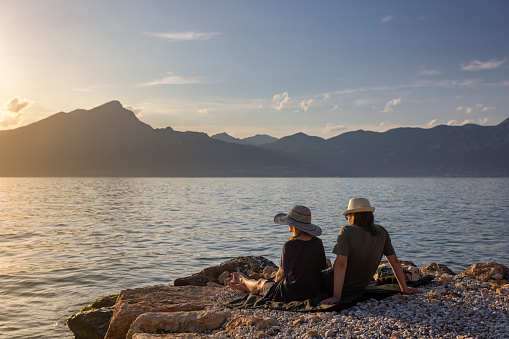 Two men hugging and leaning on a fallen log enjoy drinking mate and gazing at the lake pier and mountains on a sunny day.