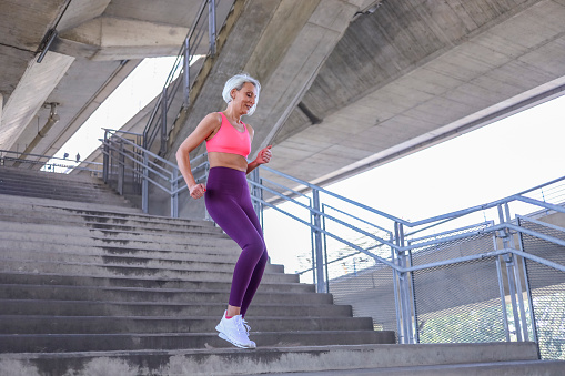 Senior woman with grey hair  in sports clothes exercising outside while listening to music. Beautiful mature woman jogging.
