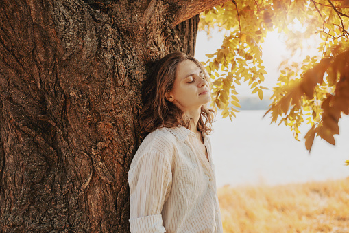 Beautiful young relaxed woman in white blouse enjoying nature while standing by a tree on an autumn day, meditating with closed eyes
