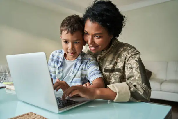 Photo of Boy sitting at the knees of his military mother and looking at the laptop screen