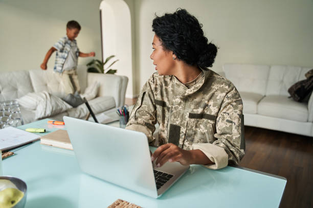istock Female soldier sitting at the table and using laptop while looking at her little son 1341245738