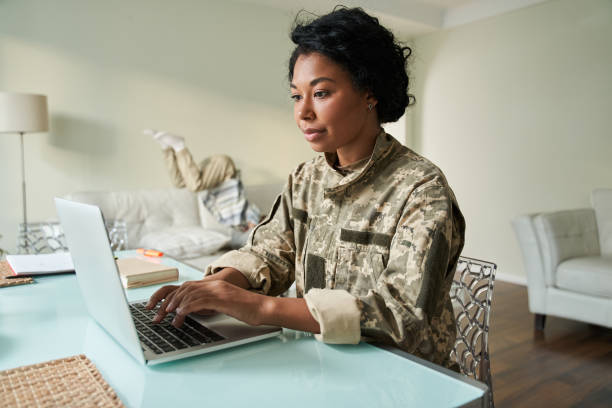 istock Female soldier sitting at the table and using laptop while her little son jumping 1341245741