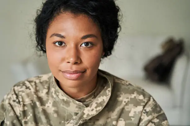 Portrait of young female at home indoor wearing green military jacket. Multiracial soldier woman wearing uniform looking at the camera while returning at home