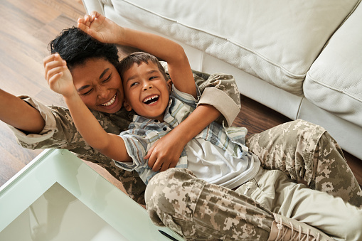 Happy black mother and her son lying on floor. Concept of family relationship and enjoying time together. Female soldier wearing camouflage uniform. After war rehabilitation. Interior of modern flat