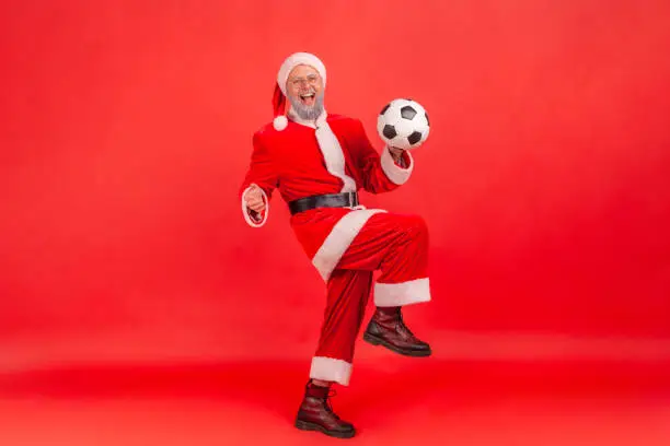Photo of Full length portrait of elderly man with gray beard wearing santa claus costume standing and playing with soccer ball, celebrating winning favorite team.