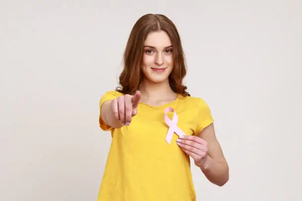 Hey you, take care of your health! Portrait of pretty optimistic teen girl in yellow T-shirt holding pink ribbon, symbol of breast cancer awareness. Indoor studio shot isolated on gray background.