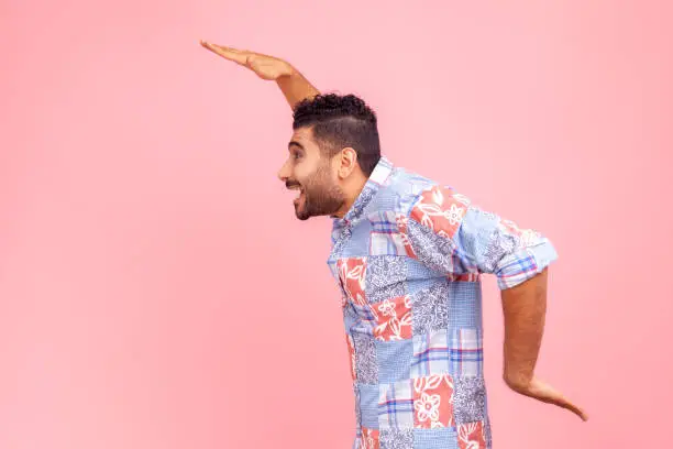 Photo of Side view of cheerful bearded man in blue casual shirt dancing, having fun and doing funny egyptian arm dance move, celebrating, rejoicing.