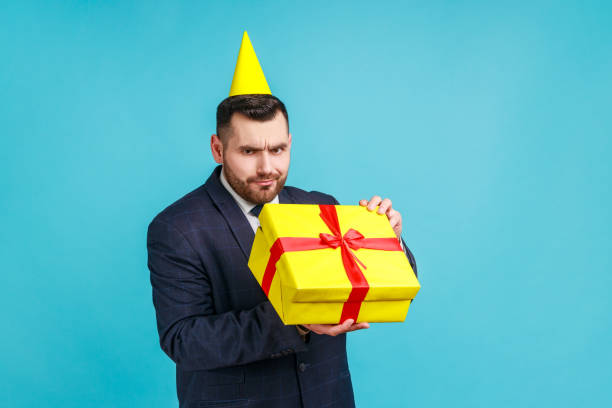 istock Unhappy man with beard wearing dark suit and party cone opening gift box and looking inside with disappointed sad expression, unwrapping bad present. 1341255460