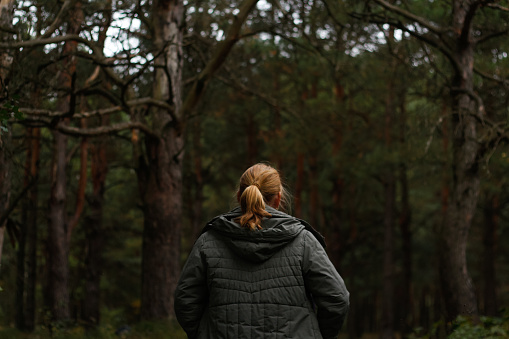 Defocus back view of a young blonde woman hiking in forest. Hiking woman walking in gloomy mystical forest, cinematic. Person discovering nature. Dark forest. Out of focus.