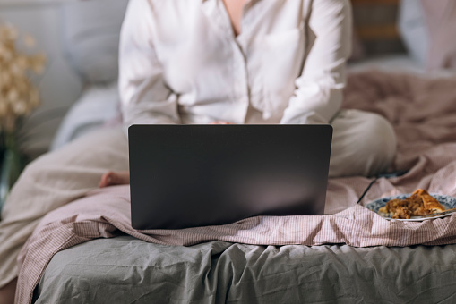 A cropped image of an unrecognisable overweight woman in pyjamas sitting on her bed, eating breakfast and working on her laptop computer