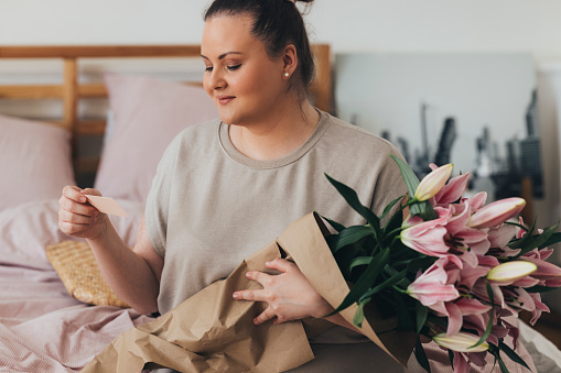 Happy young Caucasian overweight woman holding a bouquet of pink lillies and reading a card that came with them