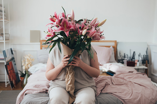 An overweight woman sitting on her bed and holding a bouquet of pink lillies
