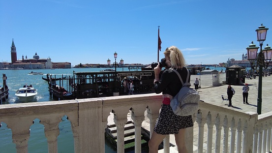 Venice, Italy - May 9, 2021: tourist woman taking pictures from venetian bridge at traditional gondola boats with tourists in tour on gondola boats in Canal of Giudecca by San Marco square.
