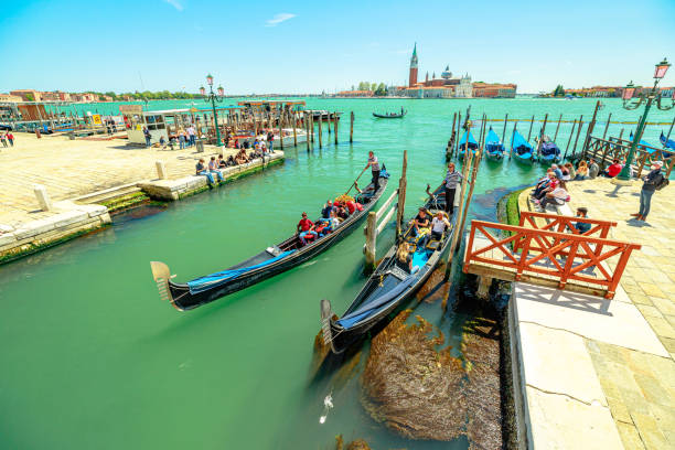 Venice, Italy - May 9, 2021: Traditional gondola boats with tourists in tour on Canal of Giudecca by Saint Mark square. San Giorgio island on background. People with mask for Coronavirus Covid19.