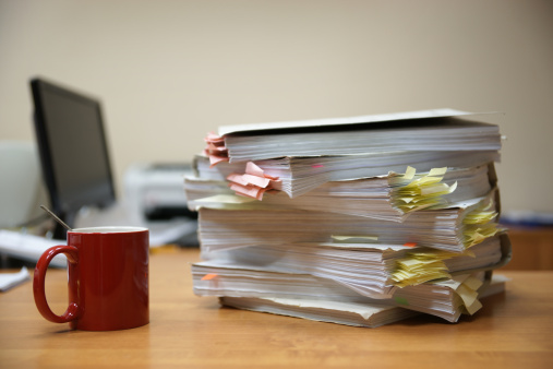 A stack of thick file folders and a cup of coffee on the office table with a monitor on background. Shallow depth of field.