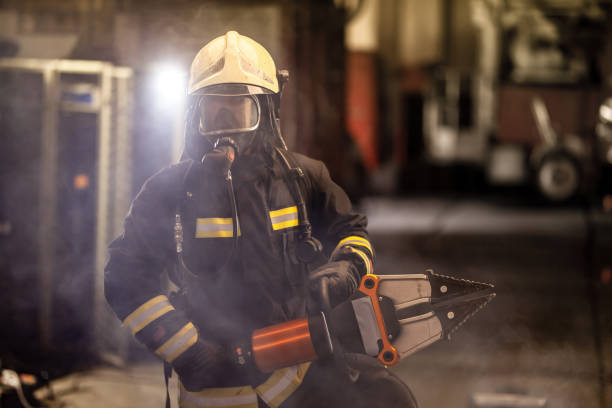 istock firefighter portrait wearing full equipment, oxygen mask, and power hydraulic cutting tool, smoke and fire trucks in the background. 1341267302
