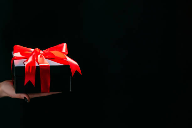 Black friday. Festive decoration. Holiday discount. Advertising background. Female hand holding black gift box red atlas ribbon isolated dark copy space.