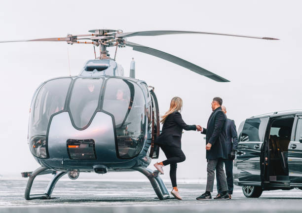 Wealthy couple traveling by a private helicopter Woman getting down the helicopter with the help from a man with their chauffeur waiting by the car on the helipad. Couple disembarking their helicopter with chauffeur standing by. helicopter stock pictures, royalty-free photos & images