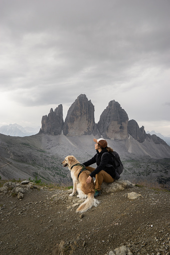 Woman hiker sitting on a rock with her pet contemplating the three peaks of Lavadero, in the Italian Alps, Dolomites.