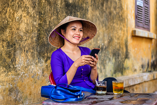 Vietnamese woman drinking traditional iced coffee - cafe sua da and using mobile phone, old town in Hoi An city, Vietnam. Cà phê sữa đá is Vietnamese iced coffee with sweetened condensed milk. Hoi An is situated on the east coast of Vietnam. Its old town is a UNESCO World Heritage Site because of its historical buildings.