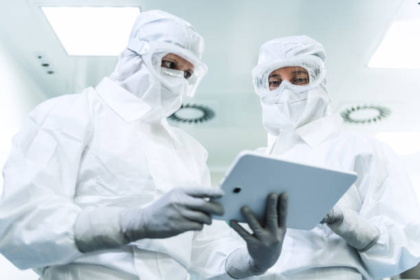 Two female researchers in protective suits Low-angle shot of two Caucasian female researchers in full protective suits, with glasses, face masks, and gloves, looking and evaluating data on the tablet. cleanroom stock pictures, royalty-free photos & images