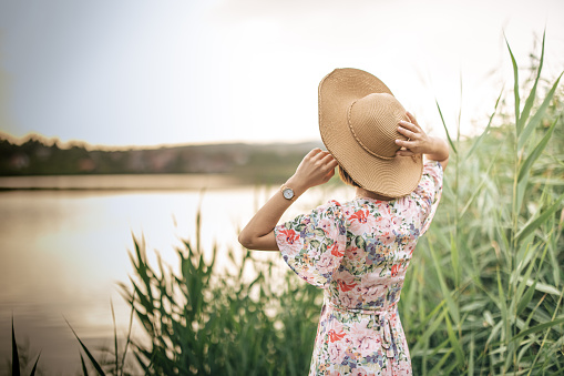 Back view of woman in dress with hat while standing on lakeshore near tall grass