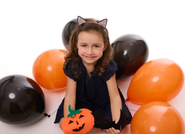 Beautiful girl child smiles with toothy smile , playing with homemade felt-cut pumpkin on the white background with lying down inflated black and orange colored balloons. Halloween concept, copy space Gorgeous girl child smiles with toothy smile , playing with homemade felt-cut pumpkin on the white background with lying down inflated black and orange colored balloons. Halloween concept, copy space black cat costume stock pictures, royalty-free photos & images
