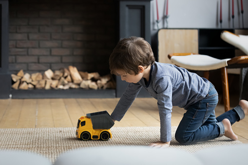 Focused boy playing with toys on heating floor at home, wheeling plastic car, trailer truck on carpet. Kid involved in game activity, improving imagination, creative skills. Childhood in safe house