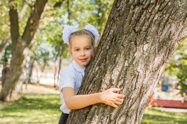 una linda colegiala con lazos blancos se para junto a un árbol en un soleado parque de otoño. primer grado el 1 de septiembre. concepto de vuelta al cole - portrait little girls 8 9 months hair bow fotografías e imágenes de stock