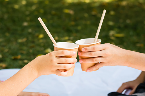 Two children's hands are holding eco-friendly cups and tubes with apple juice at a picnic in the park. Eco-friendly disposable tableware