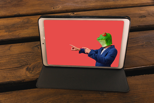 tablet on a wooden table in a garden showing a man with a frog mask on a red background with copy space