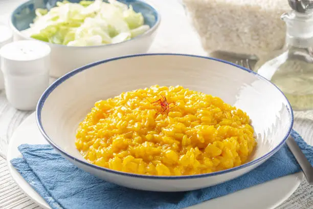 Italian dinner with risotto alla milanese and fresh salad. Italian dish made from saffron, rice, butter, hard cheese and vegetable broth. Raw arborio rice on background. White table.